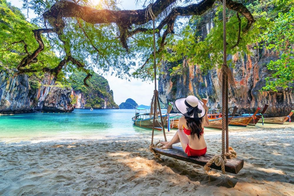 A swing hanging from a tree on a Hong Islands beach with longtail boats moored in turquoise shallows
