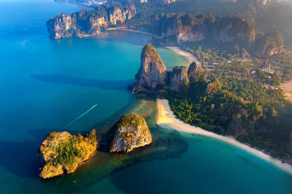 The Railay peninsula from the air: limestone cliffs enclosing a curved beach, longtail boats moored in green water