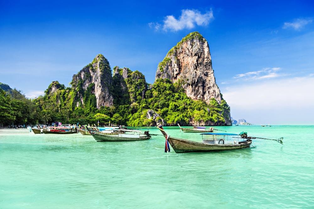 Longtail boats moored in shallow turquoise water beneath a sheer limestone wall at Railay