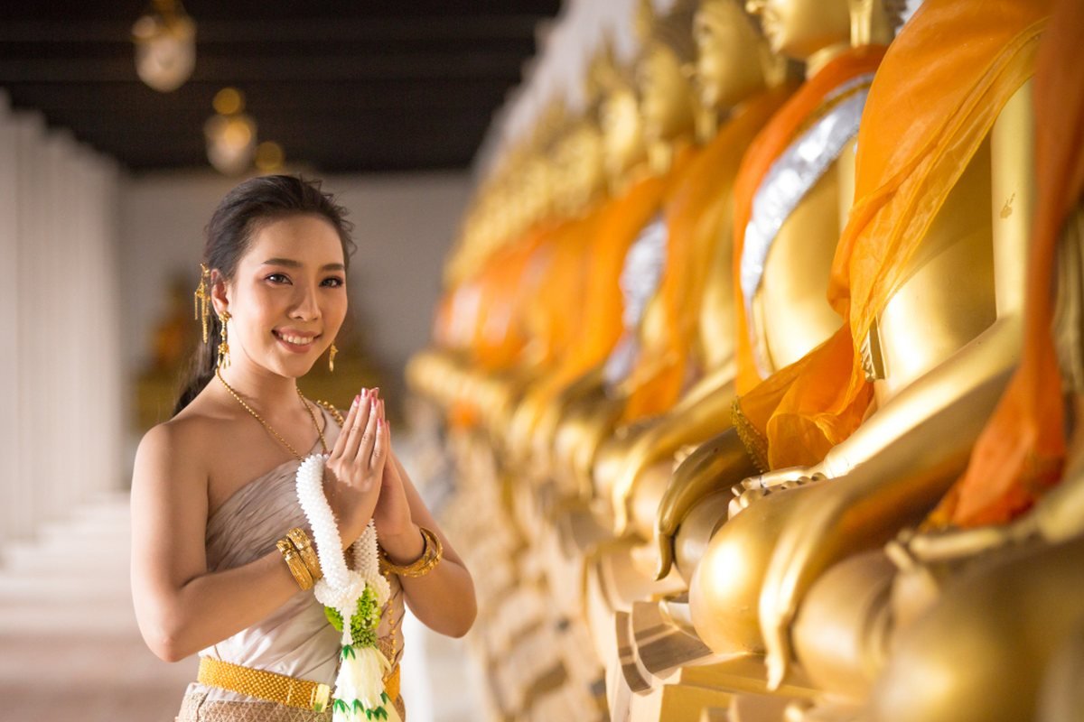 A Thai welcome, hands together in a wai, before a row of gilded Buddha images in a temple cloister